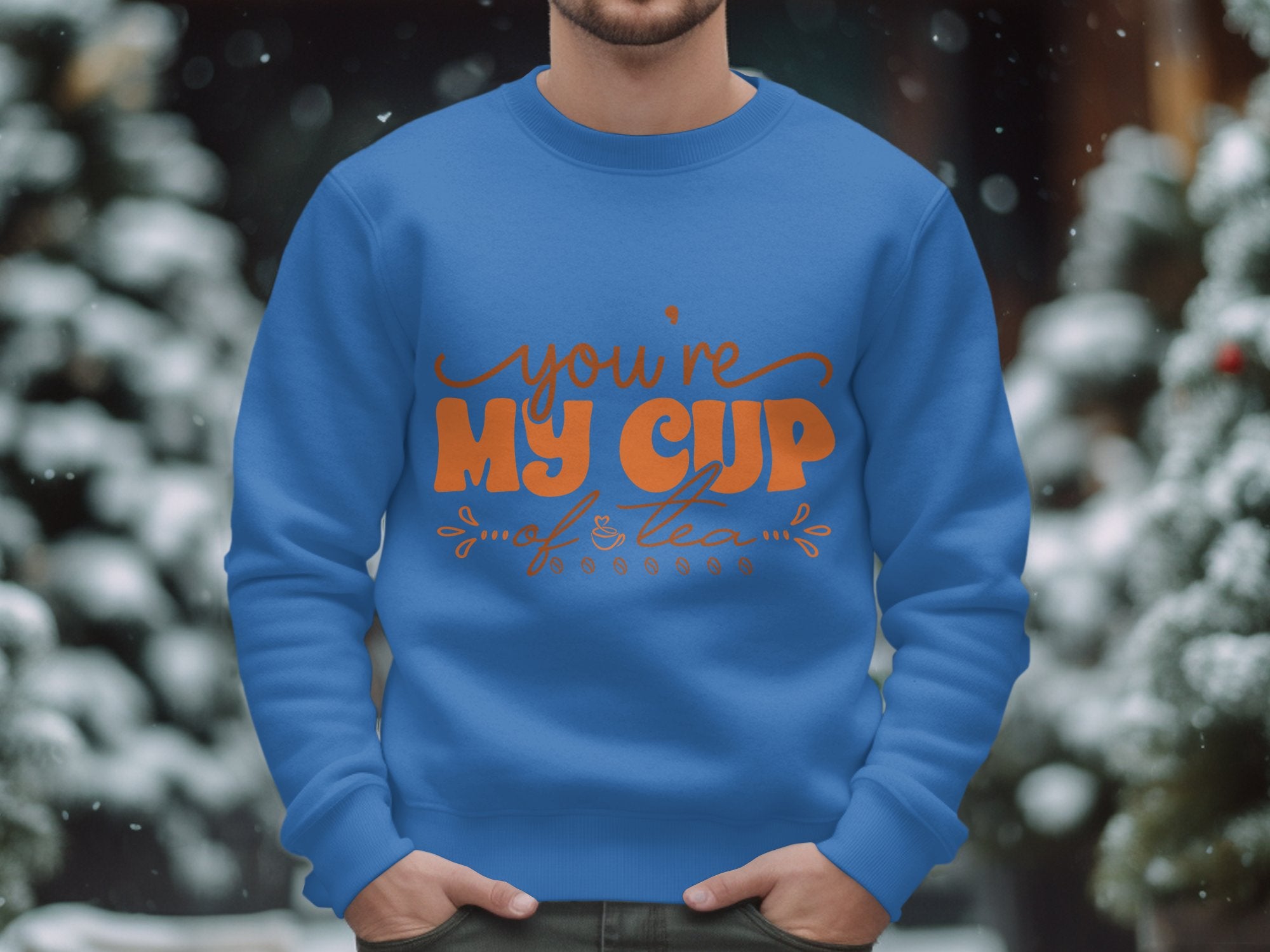 A person wearing a cozy Garment Graphics sweatshirt featuring Youre My Cup of Tea printed in orange stands against a backdrop of blurred, snow-covered trees.