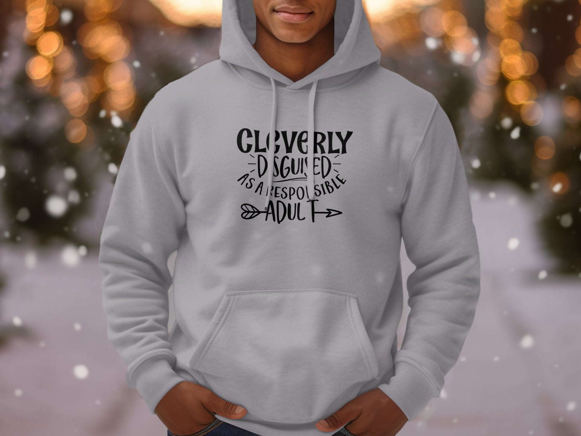 The person sports a gray Garment Graphics Cleverly Disguised As A Responsible Adult hoodie amidst a snowy, light-speckled backdrop. The blurred lights and falling snowflakes perfectly complement the playful tone of the hoodie.