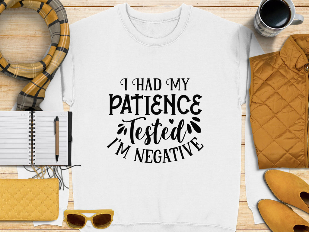 A white Garment Graphics t-shirt with I had my patience tested, Im negative text lays on a wooden surface. Ideal for casual outings, its surrounded by a notebook, pen, quilted items in yellow and orange, sunglasses, scarf, and coffee cup. Its unisex sizing ensures universal appeal.