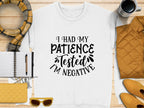 A white Garment Graphics t-shirt with I had my patience tested, Im negative text lays on a wooden surface. Ideal for casual outings, its surrounded by a notebook, pen, quilted items in yellow and orange, sunglasses, scarf, and coffee cup. Its unisex sizing ensures universal appeal.
