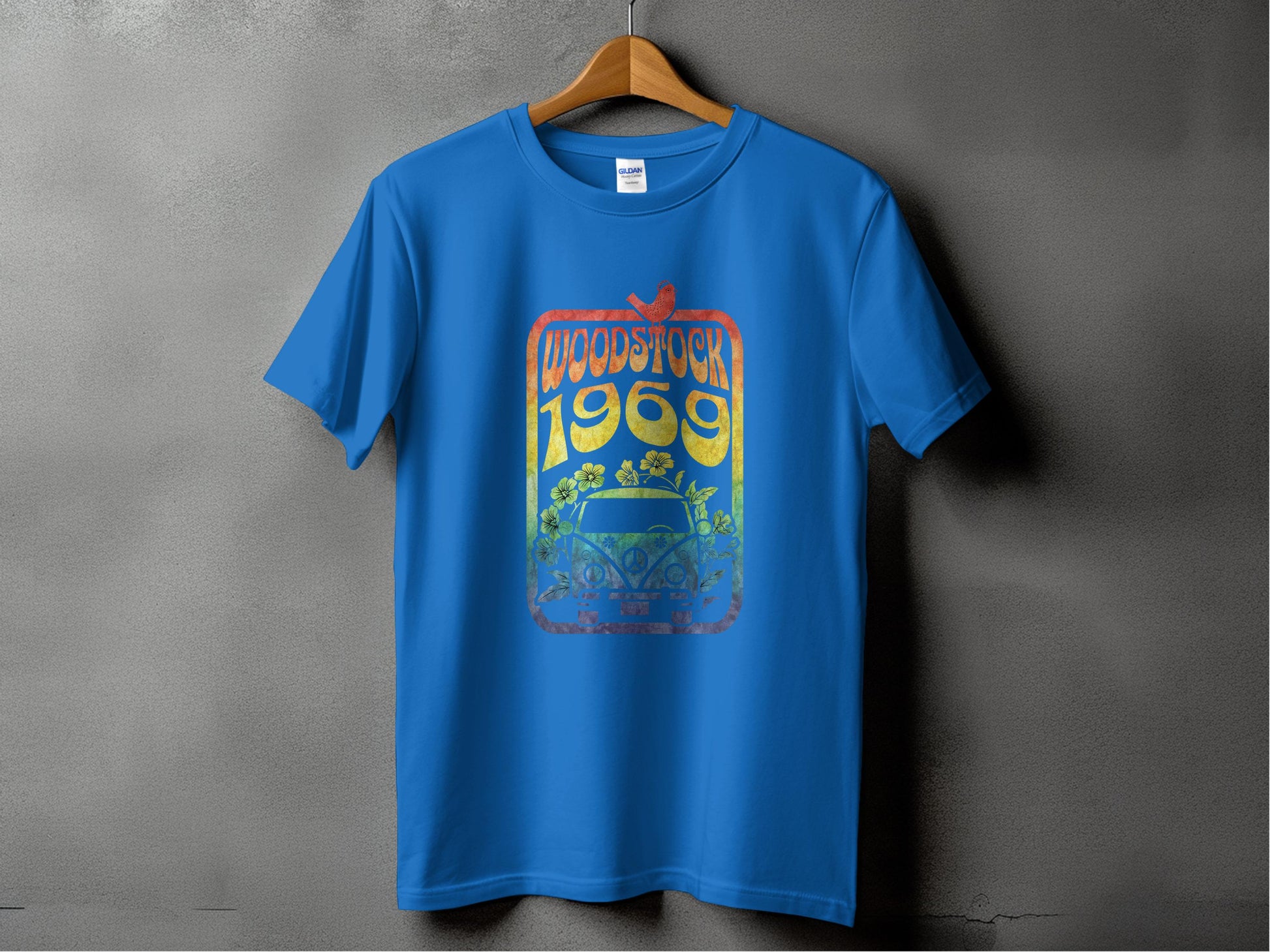 The Garment Graphics Blue T-shirt features a retro Woodstock 1969 design with a vintage car and floral elements, perfect for music lovers. Displayed on a hanger, it stands against a plain gray textured wall.