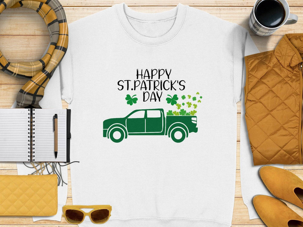 Flat lay showcasing a Garment Graphics white T-shirt with a green truck design and Happy St. Patricks Day shamrocks, encircled by festive items: yellow jacket, sunglasses, scarf, coffee cup, notebook, pen, and yellow purse on wooden surface.