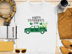 Flat lay showcasing a Garment Graphics white T-shirt with a green truck design and Happy St. Patricks Day shamrocks, encircled by festive items: yellow jacket, sunglasses, scarf, coffee cup, notebook, pen, and yellow purse on wooden surface.
