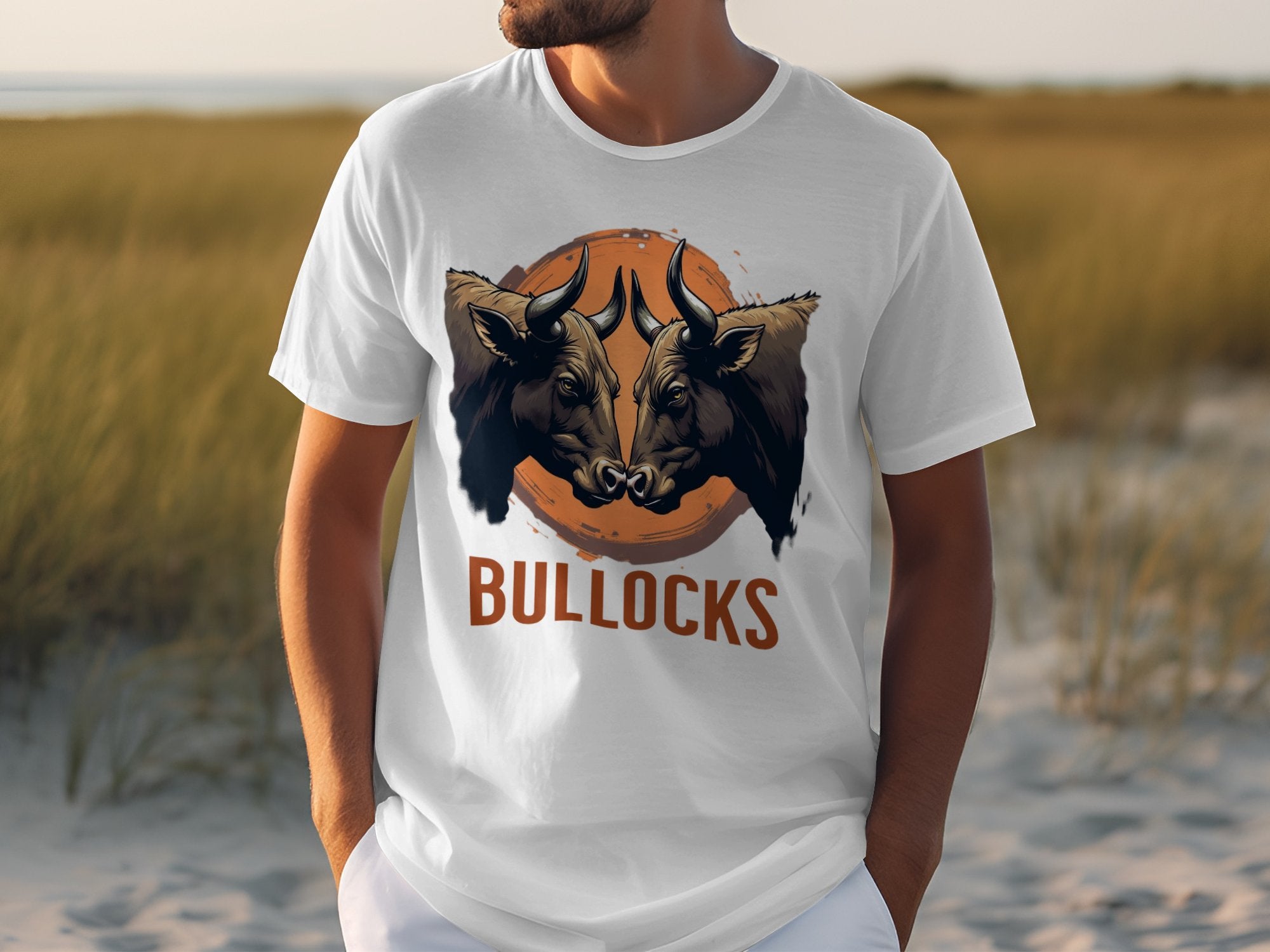 A person wears a white Garment Graphics t-shirt with two bulls facing each other in an orange circle, BULLOCKS printed below. The classic fit t-shirt is showcased against a sandy, grassy backdrop, highlighting its versatility and family sizing.