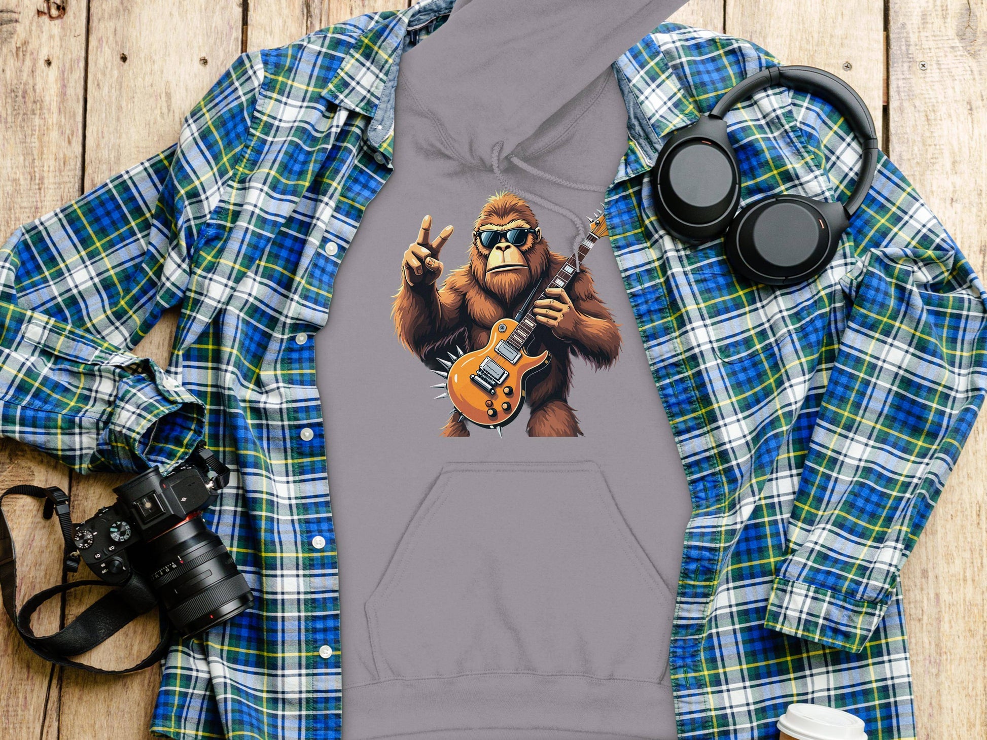 A stylish gray hoodie from Garment Graphics features a gorilla playing an electric guitar while flashing the peace sign. Laid on a blue and green plaid shirt, its perfect with a camera, headphones, and to-go coffee cup nearby—ideal for music lovers.