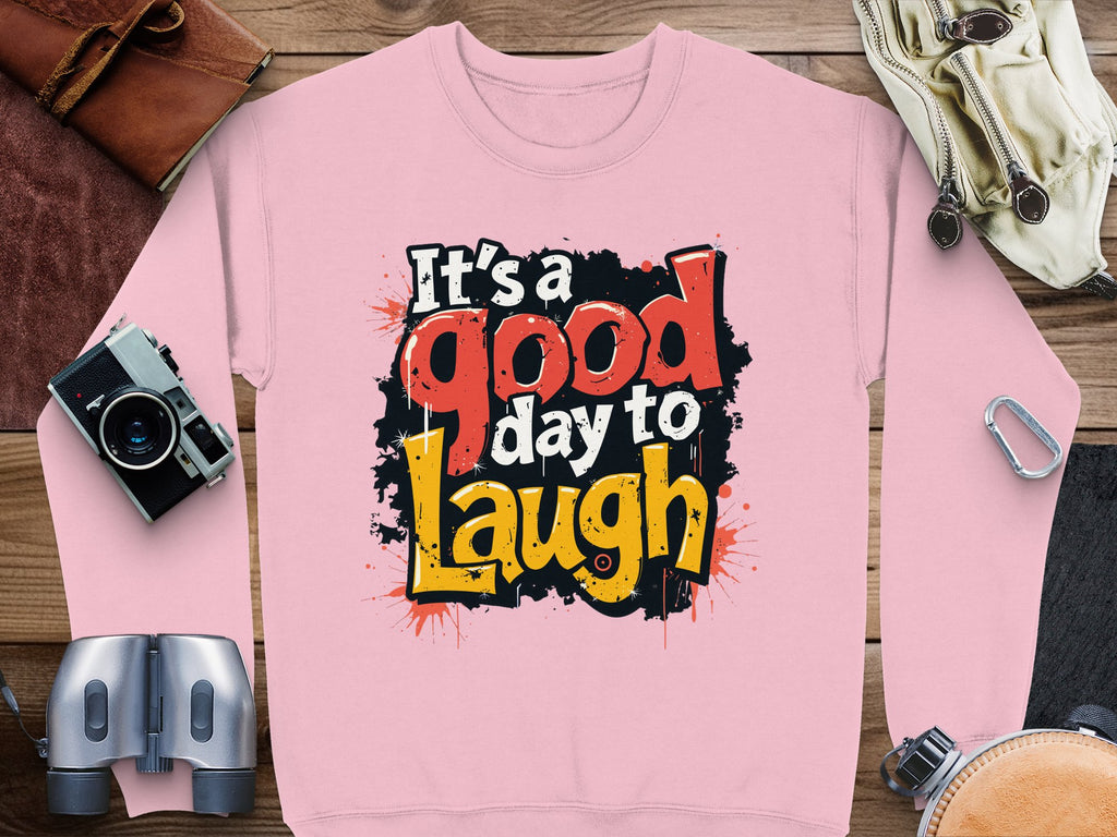 Garment Graphics presents a unisex pink sweatshirt with Its a good day to laugh in bold, colorful letters, complemented by adventure-themed items like a camera and leather bags on a wooden background.