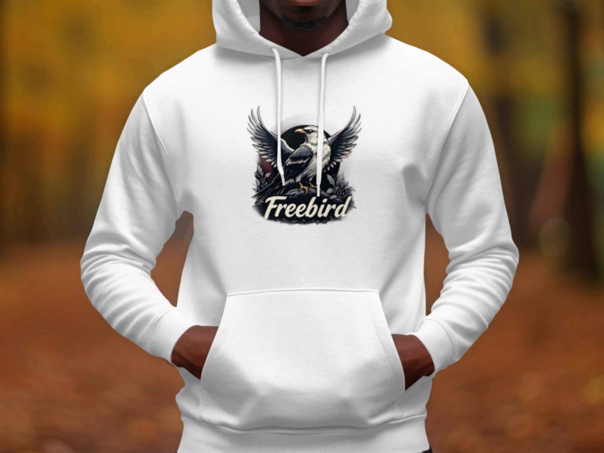 Wearing a Garment Graphics Freebird Graphic Pullover Hoodie, a person displays unique style with a bird design featuring spread wings and Freebird below, set against an autumn-hued blurred background.