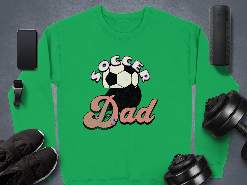 The Garment Graphics green Soccer Dad sweatshirt, with a soccer ball graphic, is set against a gray background with a smartphone, smartwatch, black shoes, dumbbells, and a wireless speaker for a retro flair.