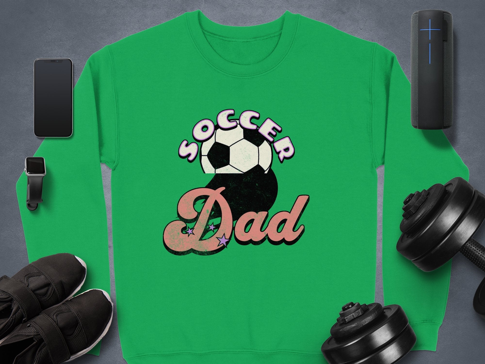The Garment Graphics green Soccer Dad sweatshirt, with a soccer ball graphic, is set against a gray background with a smartphone, smartwatch, black shoes, dumbbells, and a wireless speaker for a retro flair.