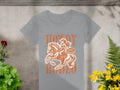 A womens light gray T-shirt from Garment Graphics features a rodeo rider graphic with Howdy Rodeo and Not Allowed in orange, set among plants and flowers.