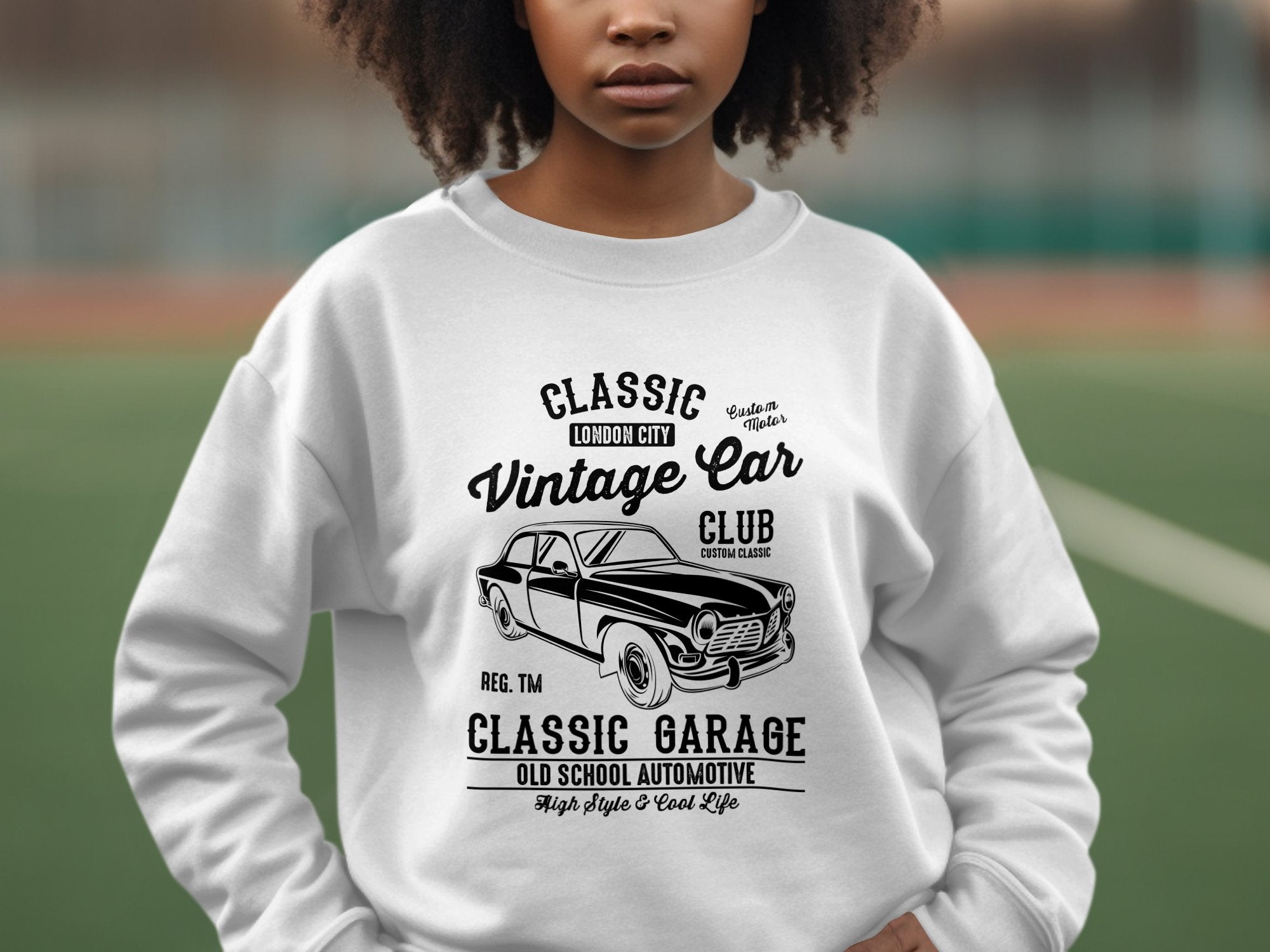 Someone wearing a Garment Graphics Vintage Car Sweatshirt featuring a black-and-white classic vehicle graphic. The text says Classic London City, Vintage Car Club, and Classic Garage Old School Automotive High Style & Cool Life, set against an outdoor sports field backdrop, embodying automotive style.