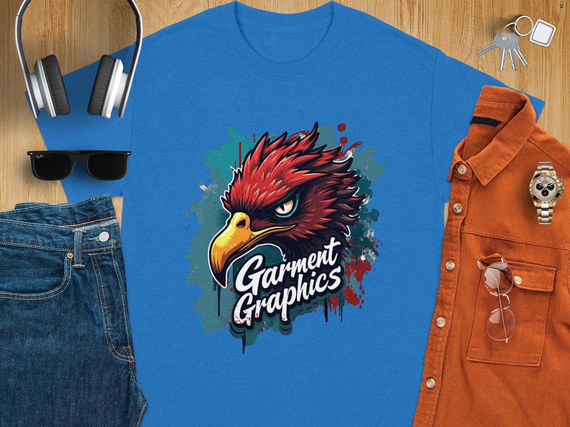 A blue Classic Fit T-shirt from Garment Graphics featuring an artistic Eagle design lies on a wooden surface, surrounded by headphones, keys, sunglasses, blue jeans, a watch, and an orange button-up shirt.