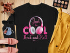 A Garment Graphics black shirt with Just be COOL, Rock and Roll in pink and white is laid on a wooden surface, ideal for music lovers. Nearby are makeup brushes, sunglasses, rock and roll headphones, a cosmetics palette, pink shoes, nail polish, and a yellow jacket.