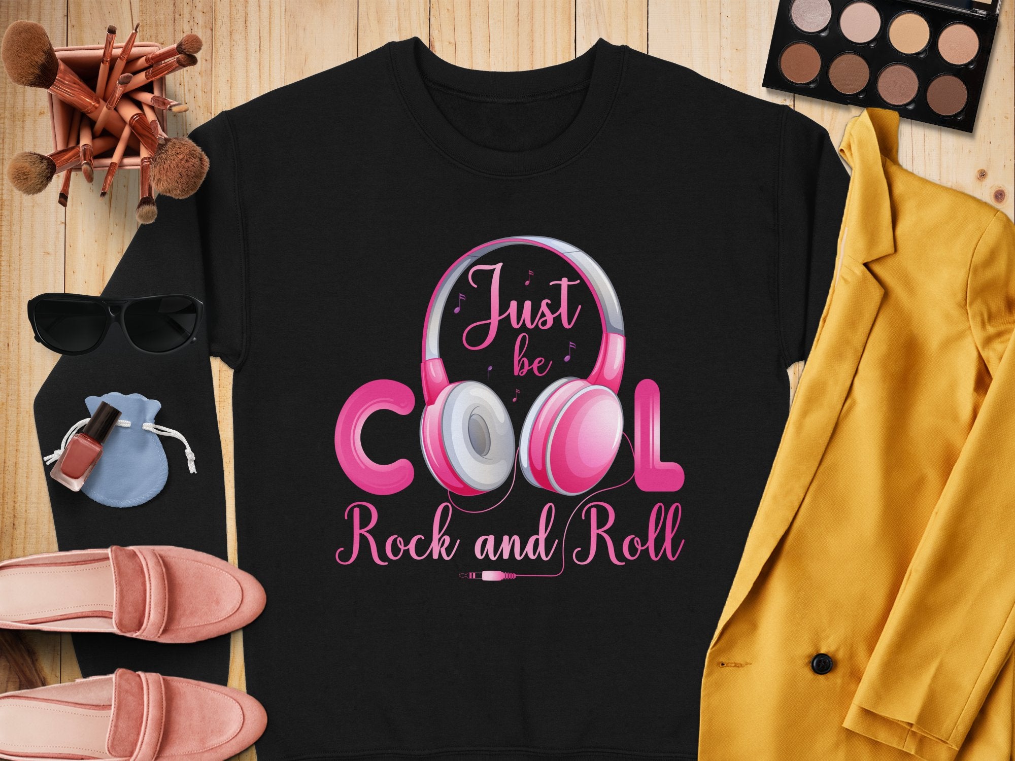 A Garment Graphics black shirt with Just be COOL, Rock and Roll in pink and white is laid on a wooden surface, ideal for music lovers. Nearby are makeup brushes, sunglasses, rock and roll headphones, a cosmetics palette, pink shoes, nail polish, and a yellow jacket.