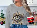 In a festive kitchen, someone holds a Santa-themed mug and wears Garment Graphics unisex graphic sweatshirt with an ornate eye design and GAZE across it, perfectly complementing the holiday decor.