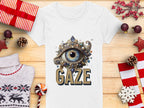 A Garment Graphics white graphic T-shirt features a jeweled eye design with GAZE lettering, set on wood. Surrounded by festive decorations like gifts, candy, and a sweater, this artistic T-shirt is available in womens sizes.