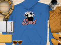 A blue hoodie from Garment Graphics features the Soccer Dad print with a soccer ball design. Its styled with vibrant yellow sunglasses, a cozy scarf, a black pen on an open notebook, a cheerful yellow purse, a steaming mug of coffee, and a quilted yellow jacket on wood.