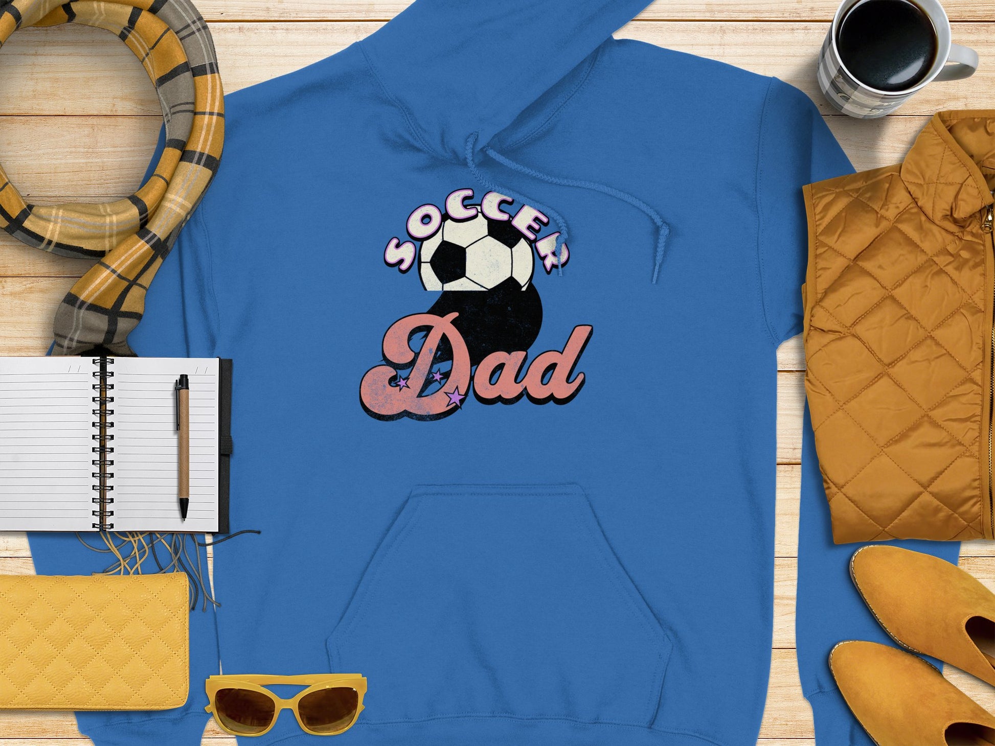 A blue hoodie from Garment Graphics features the Soccer Dad print with a soccer ball design. Its styled with vibrant yellow sunglasses, a cozy scarf, a black pen on an open notebook, a cheerful yellow purse, a steaming mug of coffee, and a quilted yellow jacket on wood.