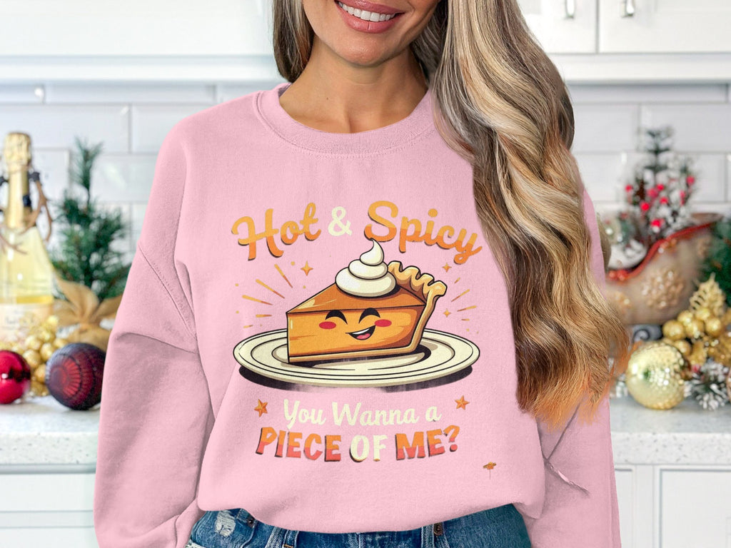A person with long, wavy hair wears a Garment Graphics pink pie graphic sweatshirt featuring a smiling pie slice with whipped cream. It reads Hot & Spicy above and You Wanna a Piece of Me? below, set against a kitchen background.