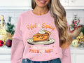 A person with long, wavy hair wears a Garment Graphics pink pie graphic sweatshirt featuring a smiling pie slice with whipped cream. It reads Hot & Spicy above and You Wanna a Piece of Me? below, set against a kitchen background.