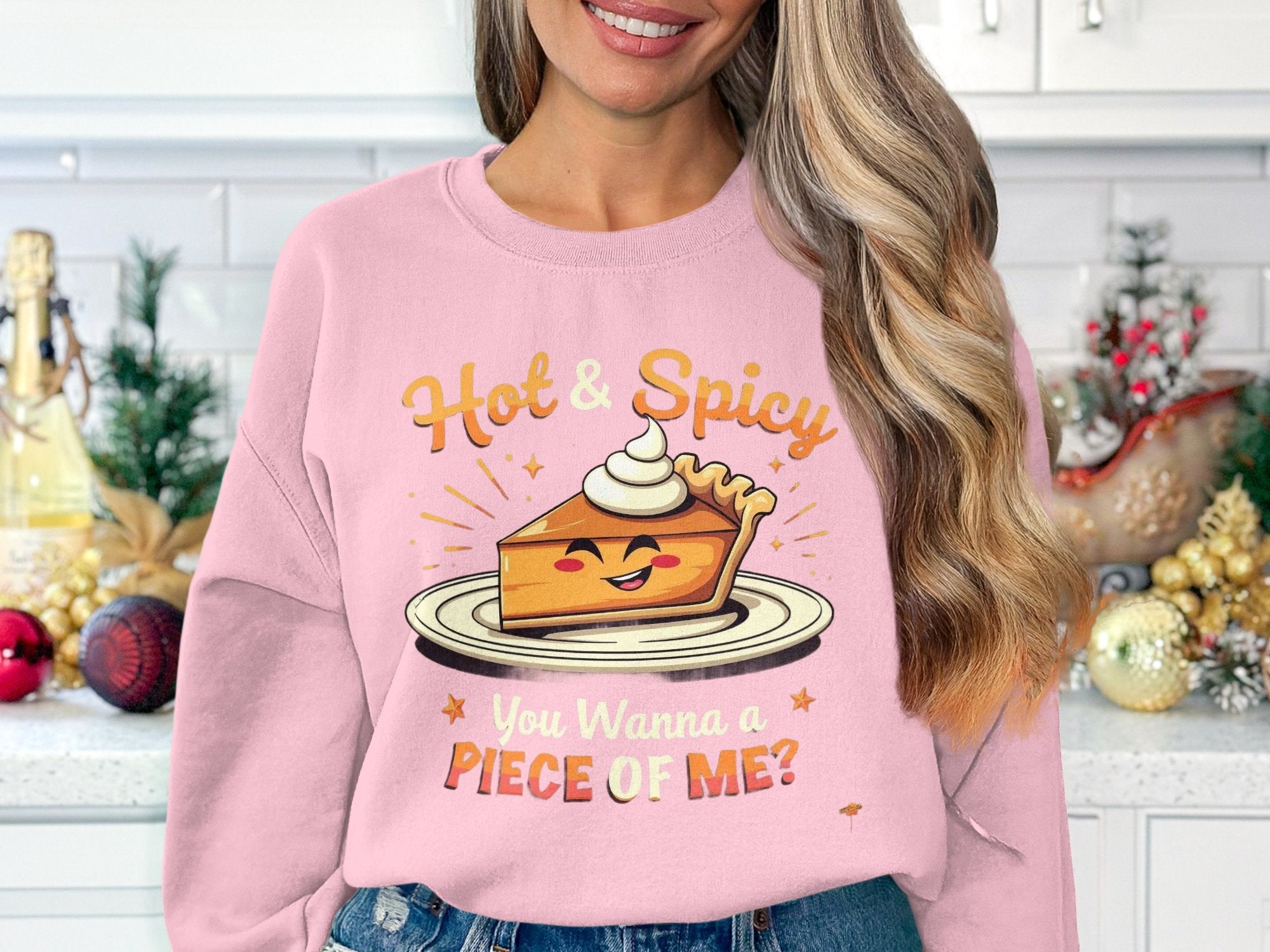 A person with long, wavy hair wears a Garment Graphics pink pie graphic sweatshirt featuring a smiling pie slice with whipped cream. It reads Hot & Spicy above and You Wanna a Piece of Me? below, set against a kitchen background.
