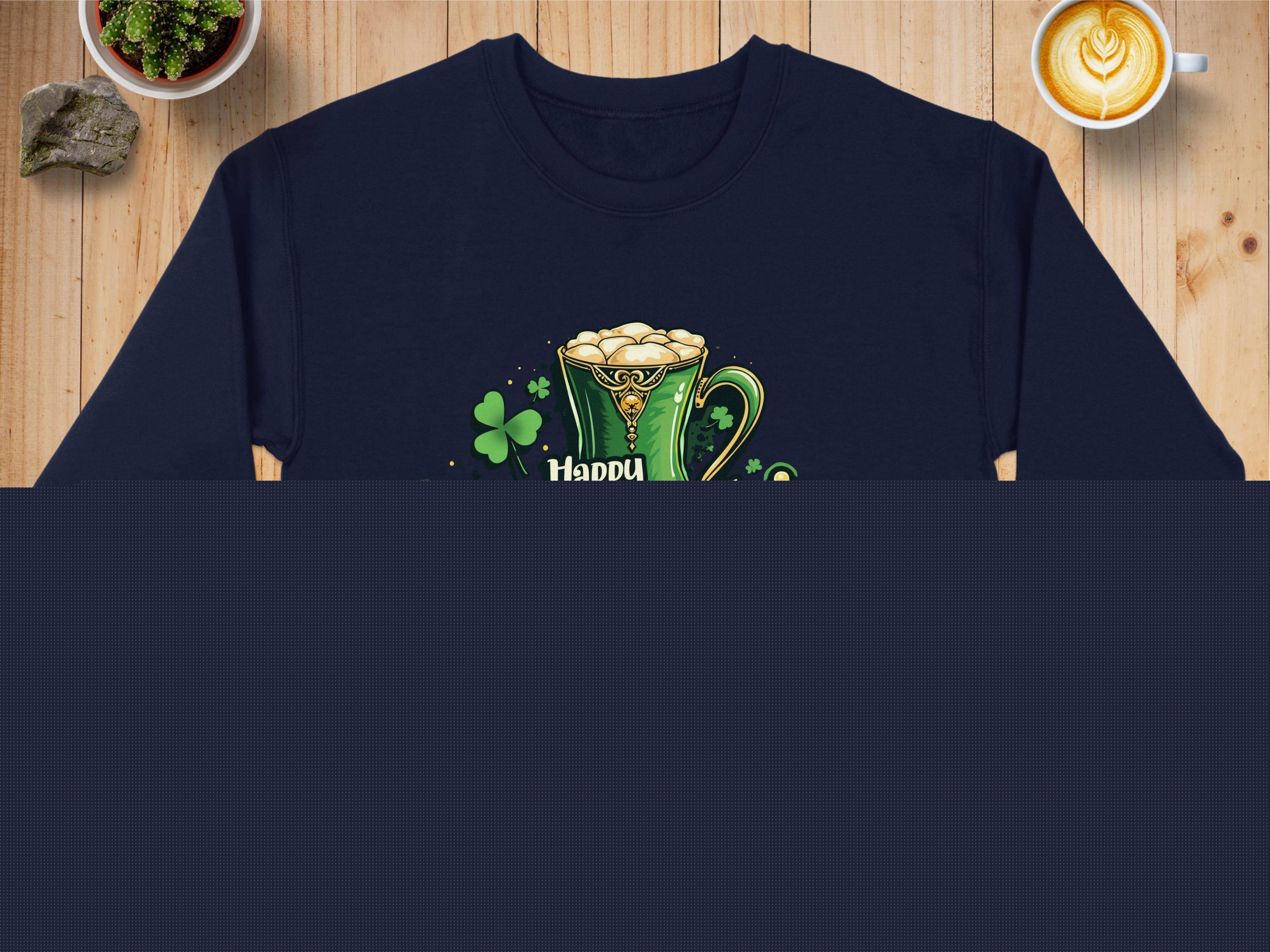 A navy blue Garment Graphics sweatshirt with a festive Happy Patricks Day graphic featuring shamrocks and a green beer mug rests on a wooden table. Nearby are a folded denim jacket, cup of coffee, and small plant completing the cozy scene.