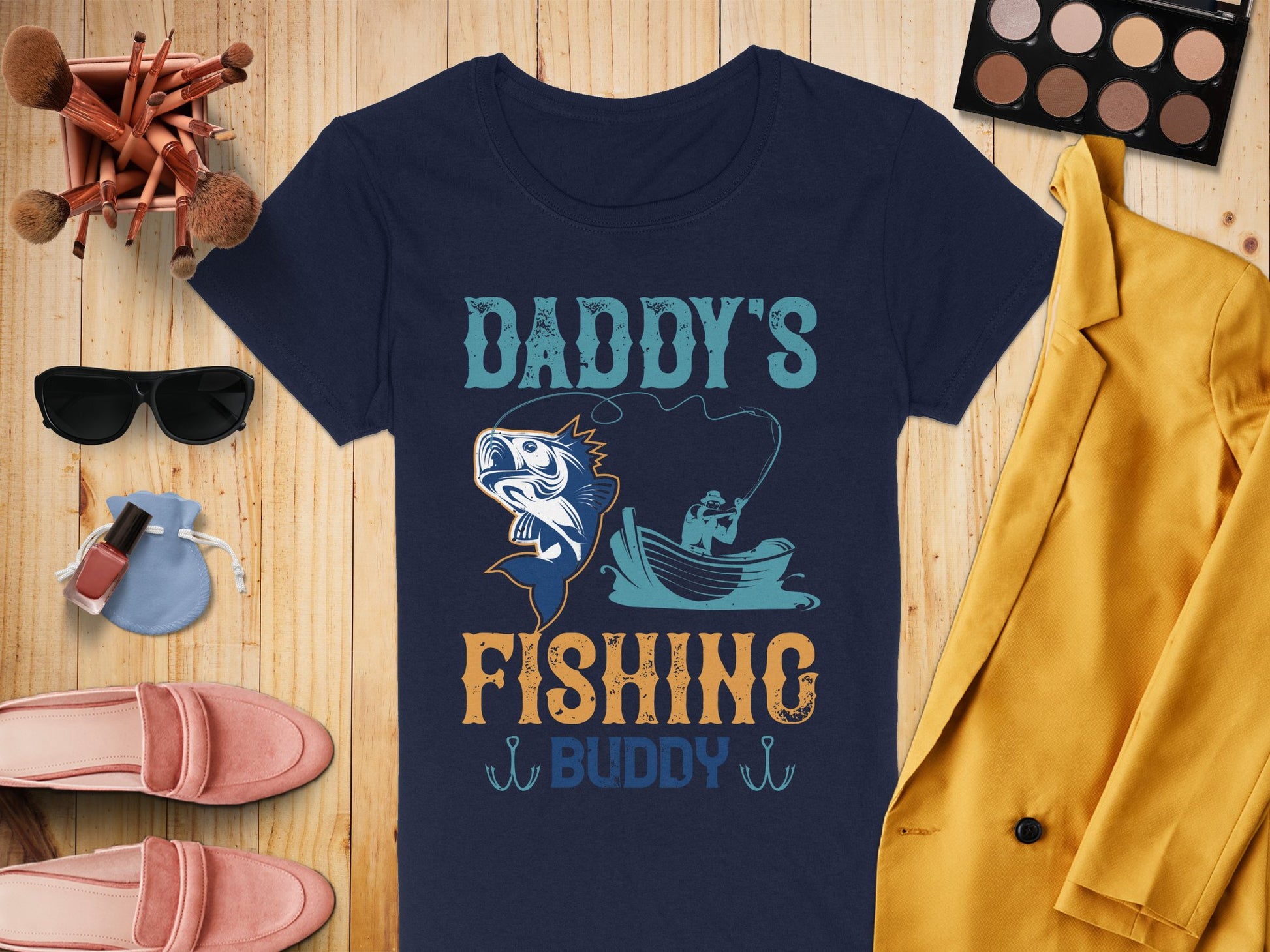 Flat lay featuring a Garment Graphics navy fishing t-shirt with Daddys Fishing Buddy and a fish graphic, surrounded by a yellow blazer, pink loafers, makeup brushes, brown eyeshadow palette, sunglasses, blue pouch, and nail polish on a wooden surface.