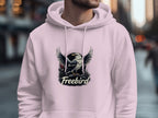 A person dons a light pink Freebird Graphic Pullover Hoodie by Garment Graphics on a city street. The eagle design adds flair, while the blurred backdrop of buildings and a passerby enhances the urban atmosphere.