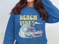 A person is wearing a Garment Graphics blue sweatshirt featuring a graphic design of a classic car with surfboards, set against waves, palm trees, and the sun. The text Beach Vibes perfectly captures retro beach fun vibes.