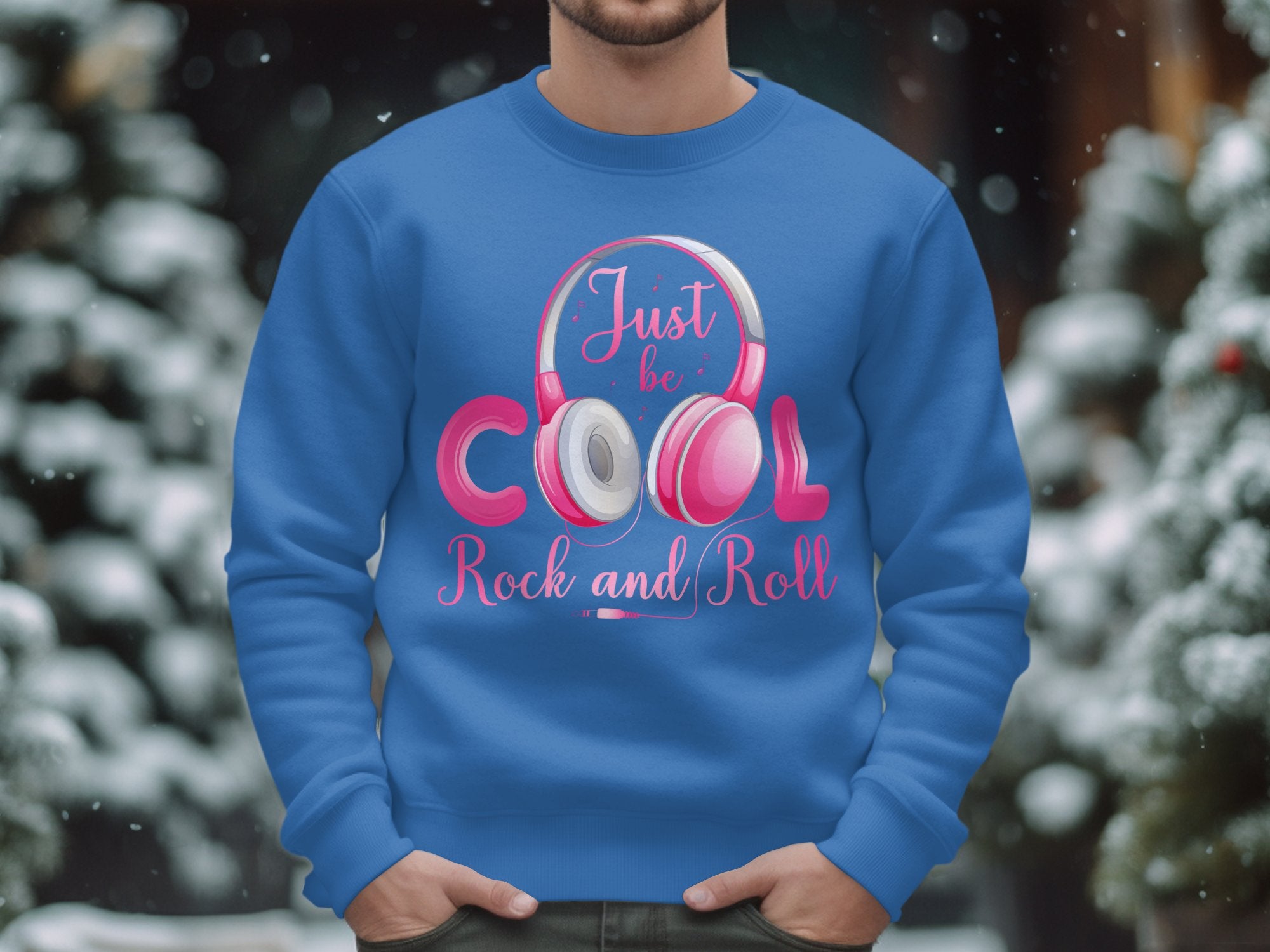 Someone is wearing a trendy Garment Graphics blue sweatshirt with “Just be Cool Rock and Roll” in pink and white and headphone graphics, set against snowy trees—a perfect winter statement for stylish music lovers.