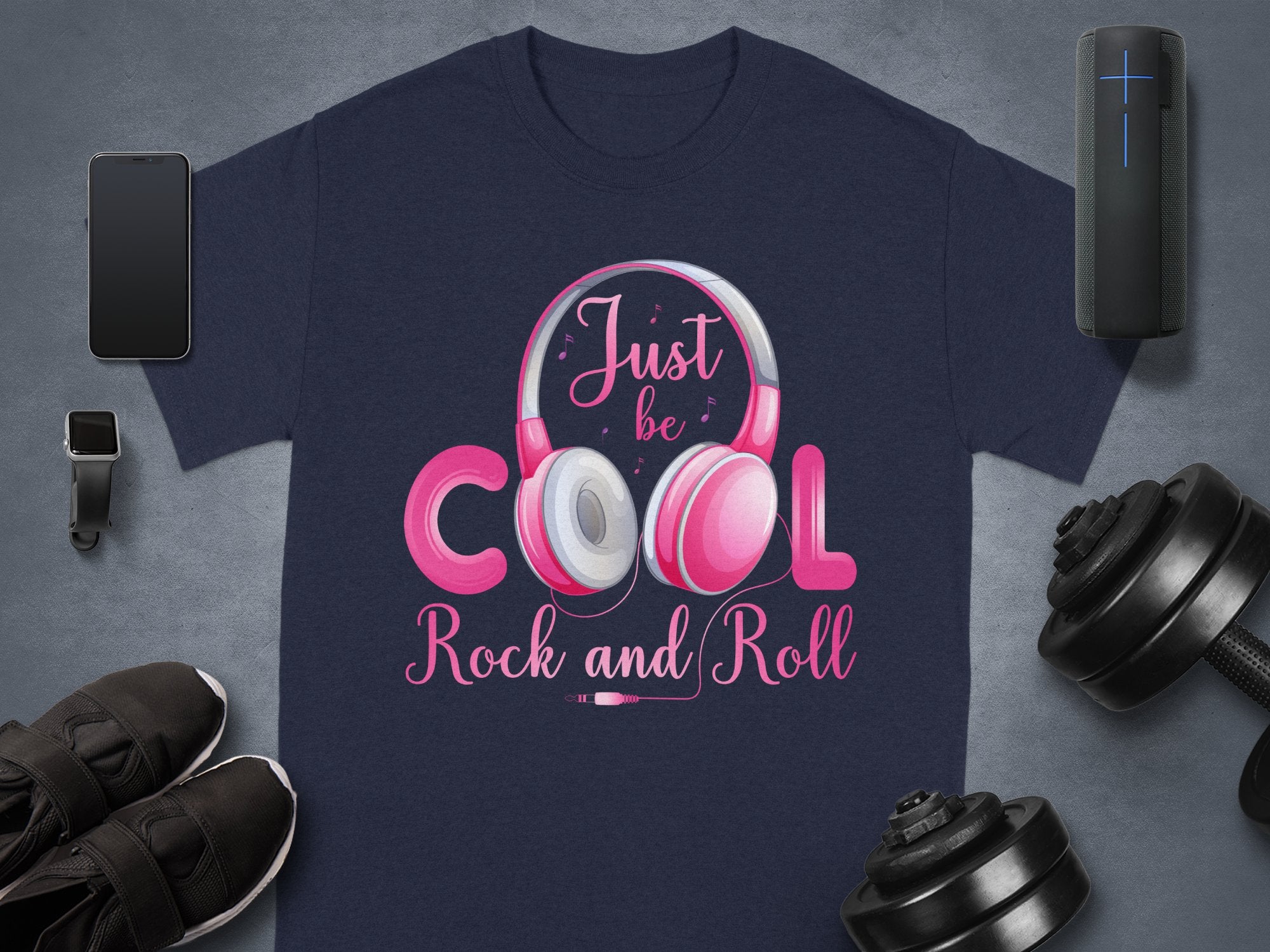 A navy blue T-shirt by Garment Graphics features pink and white lettering with a fun headphones graphic that reads Just be cool Rock and Roll. Ideal for music lovers, it pairs perfectly with a smartwatch, smartphone, black sneakers, wireless speaker, and adjustable dumbbells.
