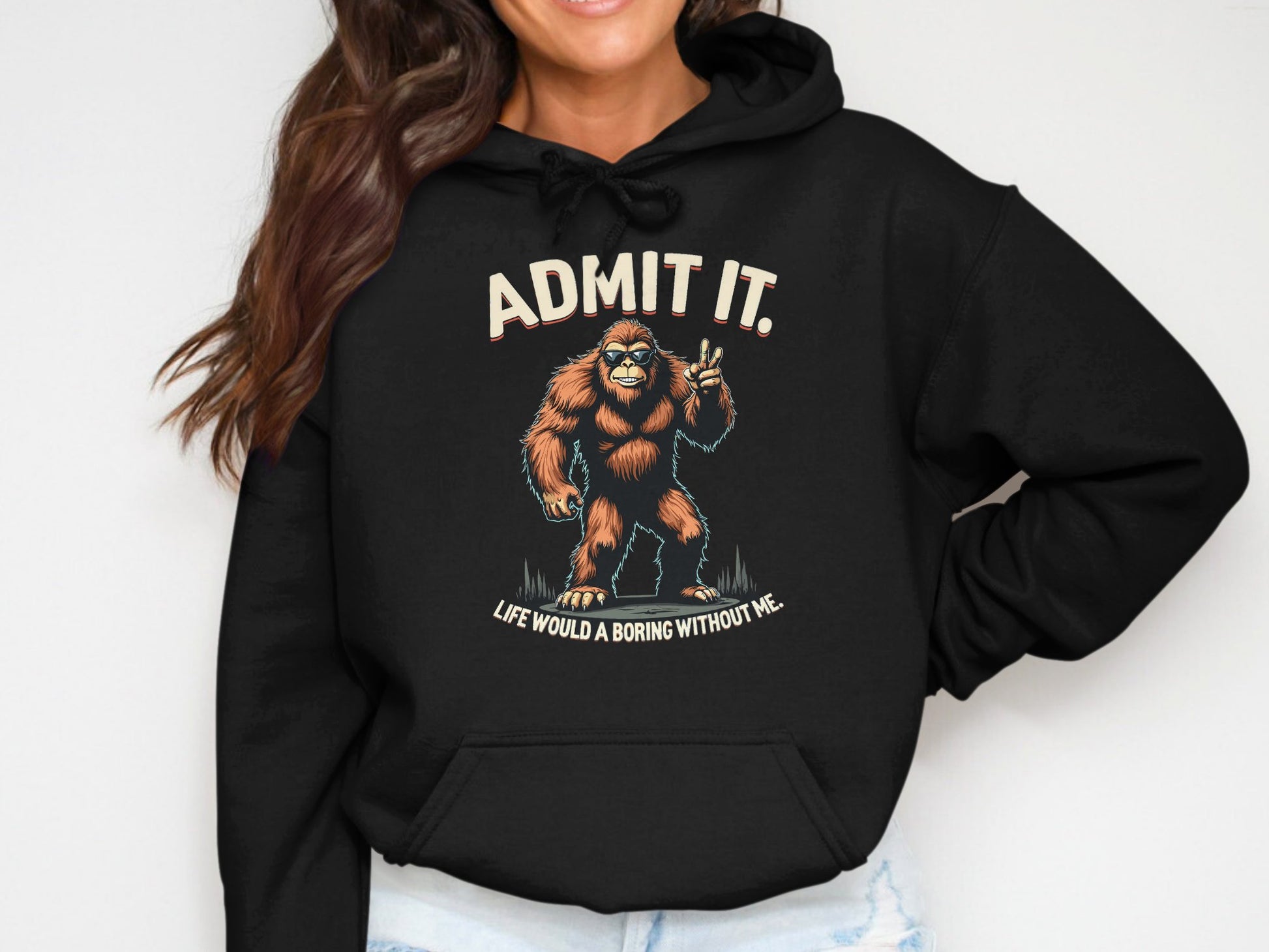 Wearing a classic fit black hoodie from Garment Graphics, featuring the phrase ADMIT IT. LIFE WOULD BE BORING WITHOUT ME and a Bigfoot illustration making a peace sign, this cool character grins beneath wavy brown hair, radiating quirky charm.