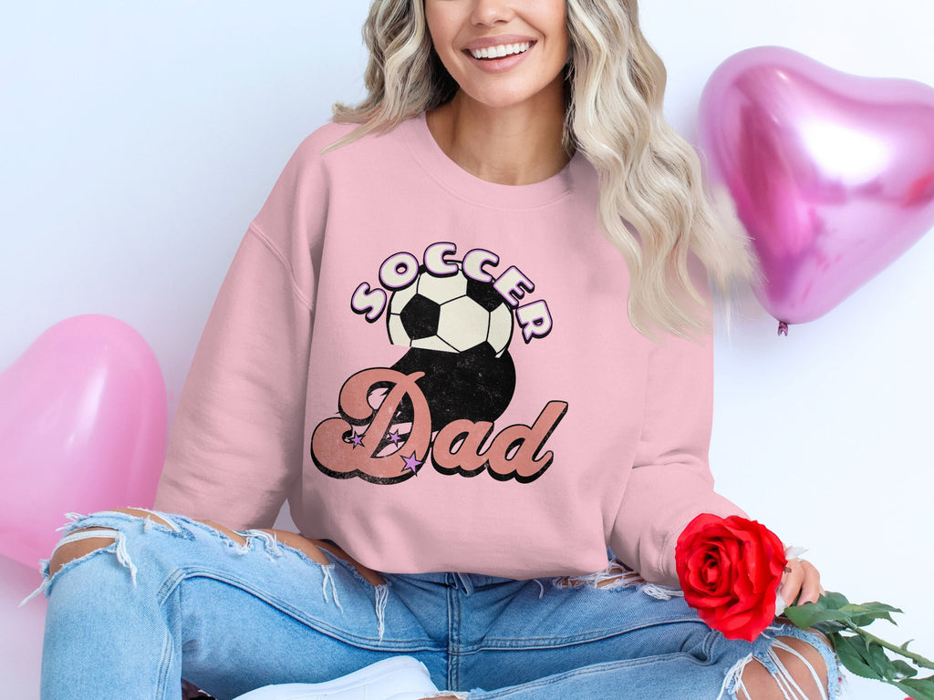 A person with long blond hair smiles in a pink Soccer Dad sweatshirt with a retro design by Garment Graphics. They sit holding a red rose, flanked by two pink heart-shaped balloons, completing the look with stylish ripped jeans.