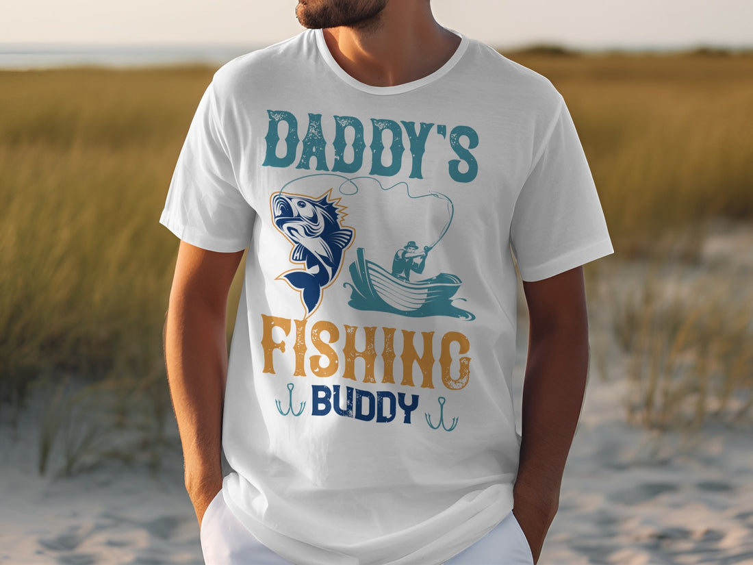 A person wears a Garment Graphics t-shirt, featuring Daddys Fishing Buddy with a fun fish and boater scene, decorative hooks, and set against a sandy background with hints of grass. Perfect for fishing fans.