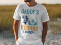 A person wears a Garment Graphics t-shirt, featuring Daddys Fishing Buddy with a fun fish and boater scene, decorative hooks, and set against a sandy background with hints of grass. Perfect for fishing fans.