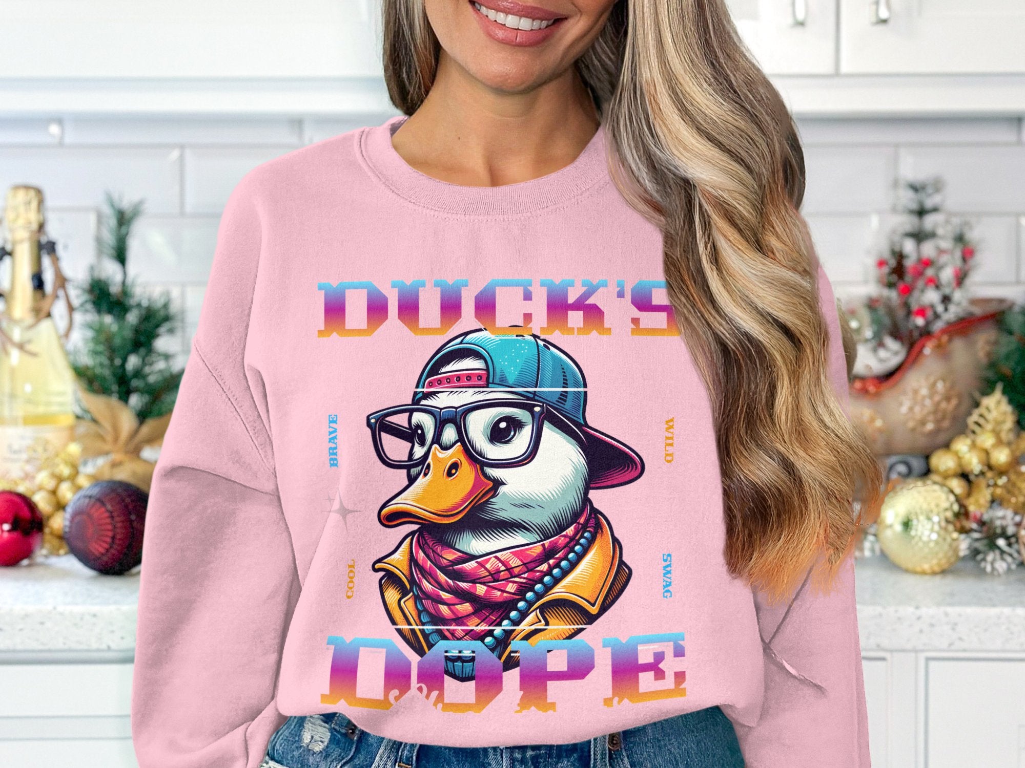 A woman boldly styles her kitchen attire with a Garment Graphics pink sweatshirt featuring a trendy cartoon duck in a cap and glasses labeled DUCKS DOPE, while blurred festive decorations adorn the countertop behind her.