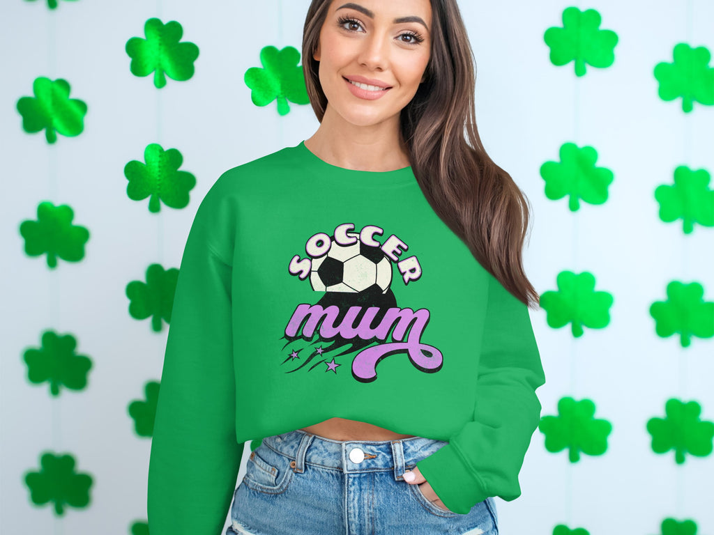 A woman with long brown hair smiles warmly in front of a white wall decorated with green shamrocks, wearing a vintage-style green Soccer Mum sweater adorned with an illustrated soccer ball. This stylish garment is from Garment Graphics.