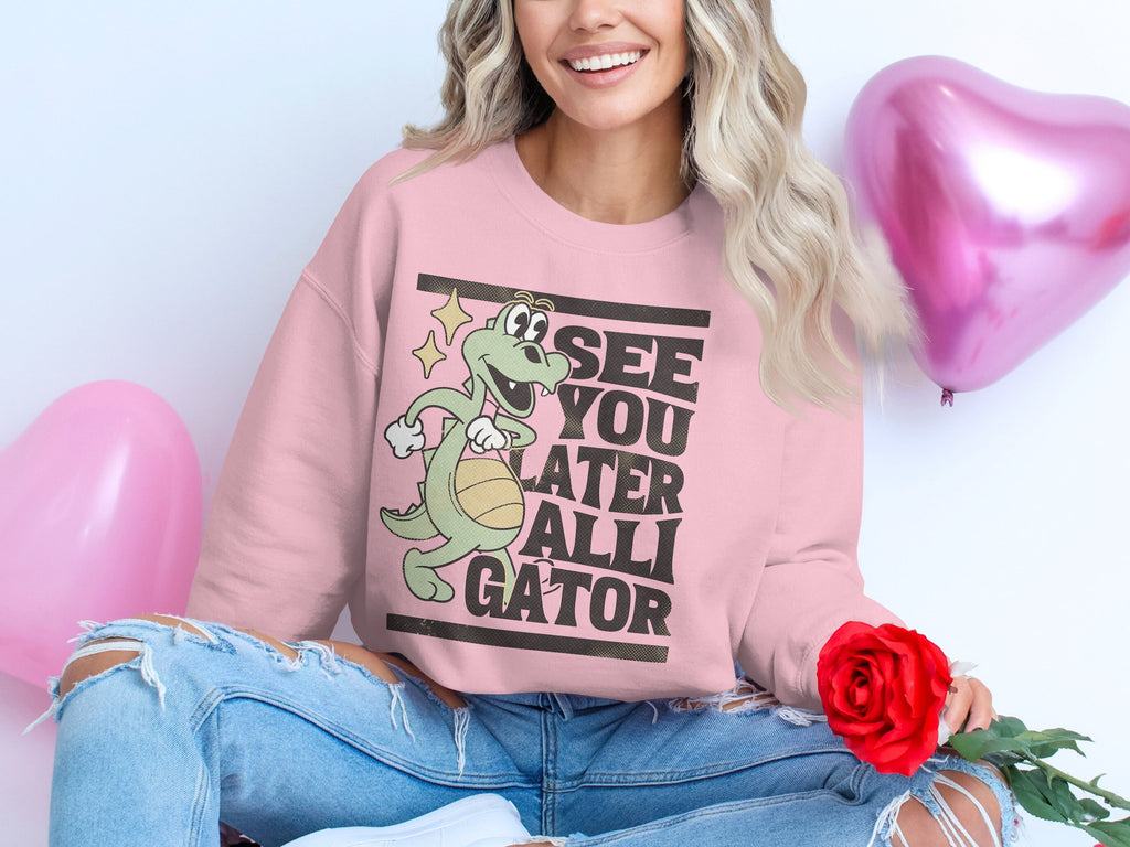 A person sits cross-legged, holding a red rose, wearing Garment Graphics pink See You Later Alligator sweatshirt with an alligator design. Heart-shaped balloons float behind them as their wavy hair falls over ripped jeans, enhancing the playful alligator graphic vibe.