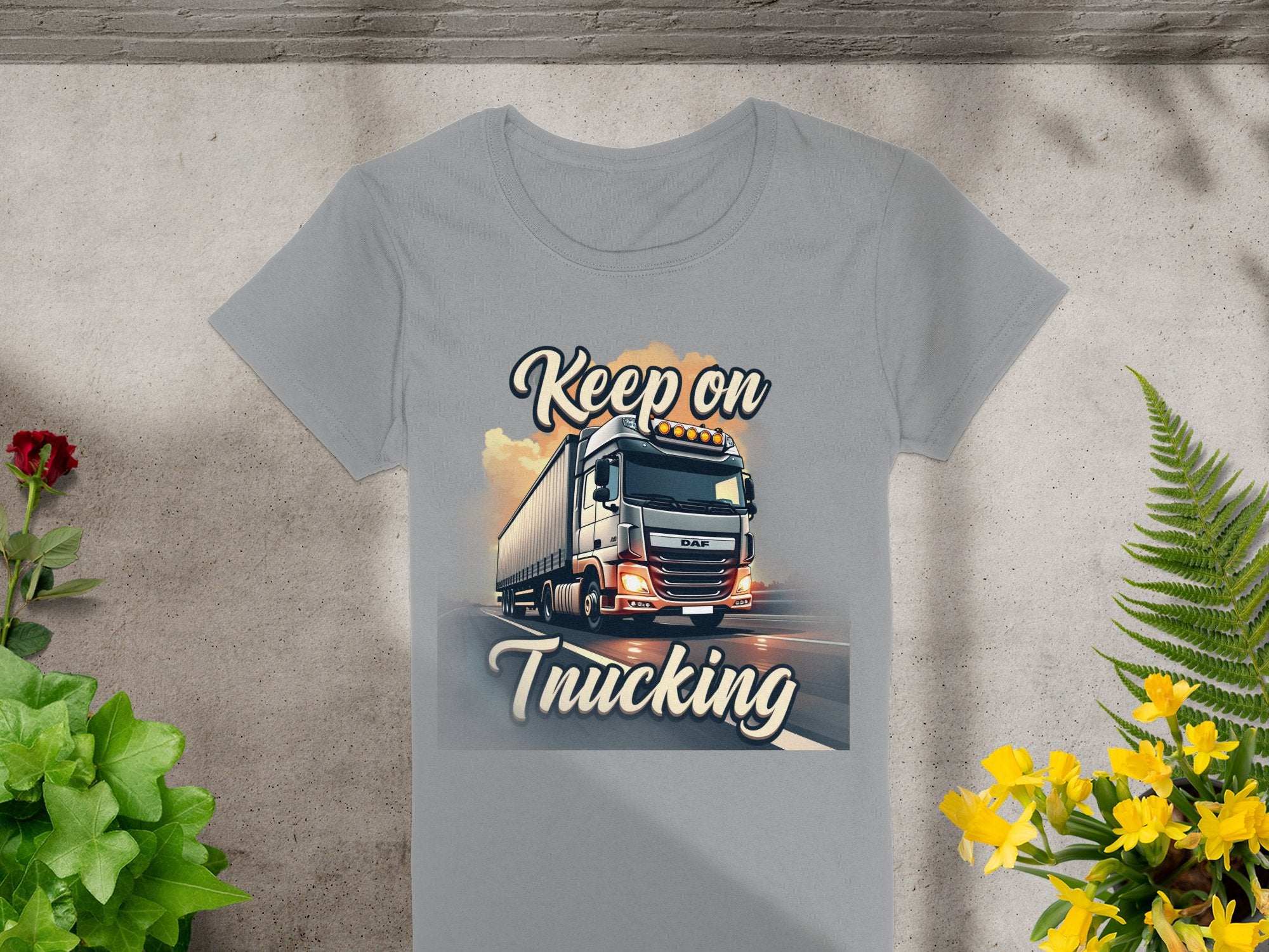 On a concrete surface, Garment Graphics gray T-shirt features a bold truck design with Keep On Trucking text. It is surrounded by small yellow flowers and green plants, making it a standout in graphic design T-shirts.