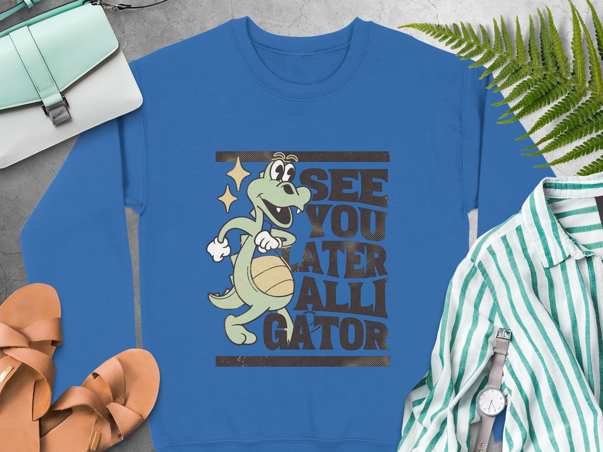 Garment Graphics presents a blue sweatshirt with a playful alligator graphic holding a basketball and the slogan SEE YOU LATER ALLIGATOR. Its styled with a green striped shirt, sandals, watch, fern leaf, and light-colored handbag on a gray surface.