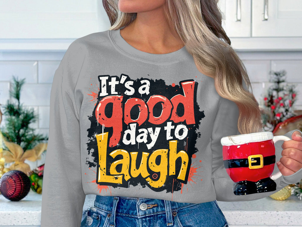 Someone holds a Santa-themed mug in the kitchen, wearing a cozy Garment Graphics hoodie that reads Its a good day to laugh in red, black, and yellow. The unisex fit offers comfort among festive decor and countertop cheer.