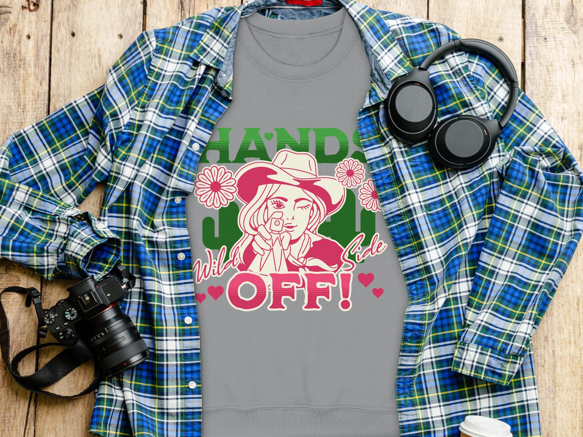 A bold Garment Graphics T-shirt on a wooden surface features a person in a hat surrounded by flowers, with Hand Off! and Wild Side text. Its displayed on a blue plaid shirt next to a camera, headphones, and coffee cup, pairing well with Wild Side Sweatshirts offering unisex sizes.