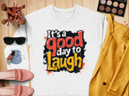 A Its a Good Day to Laugh white t-shirt by Garment Graphics, crafted from medium-heavy fabric, sits on wood. Surrounding it are a yellow jacket, pink shoes, sunglasses, makeup brushes and palette, plus a small purse—ideal for those who appreciate comfort and style!.