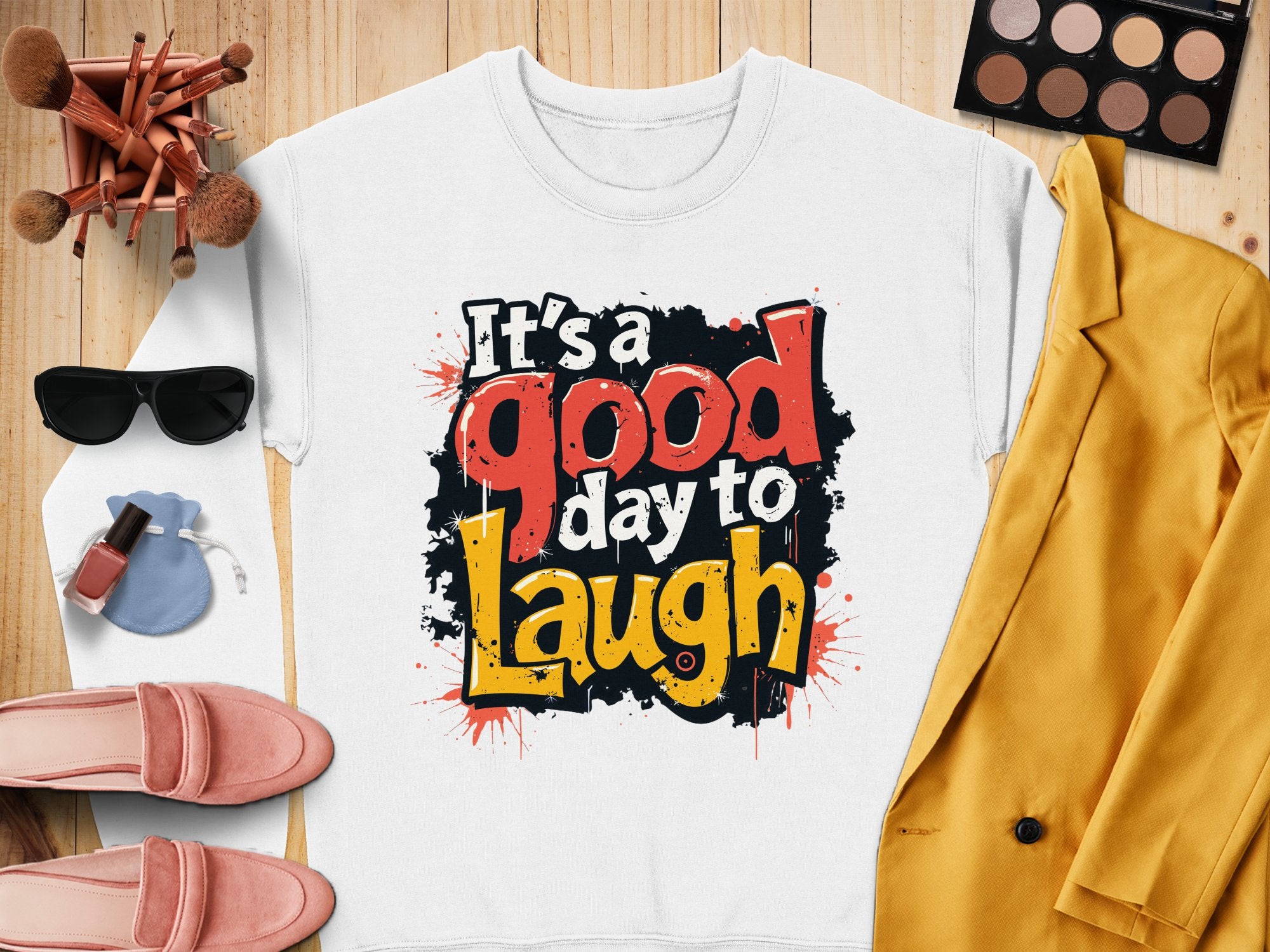 A Its a Good Day to Laugh white t-shirt by Garment Graphics, crafted from medium-heavy fabric, sits on wood. Surrounding it are a yellow jacket, pink shoes, sunglasses, makeup brushes and palette, plus a small purse—ideal for those who appreciate comfort and style!.
