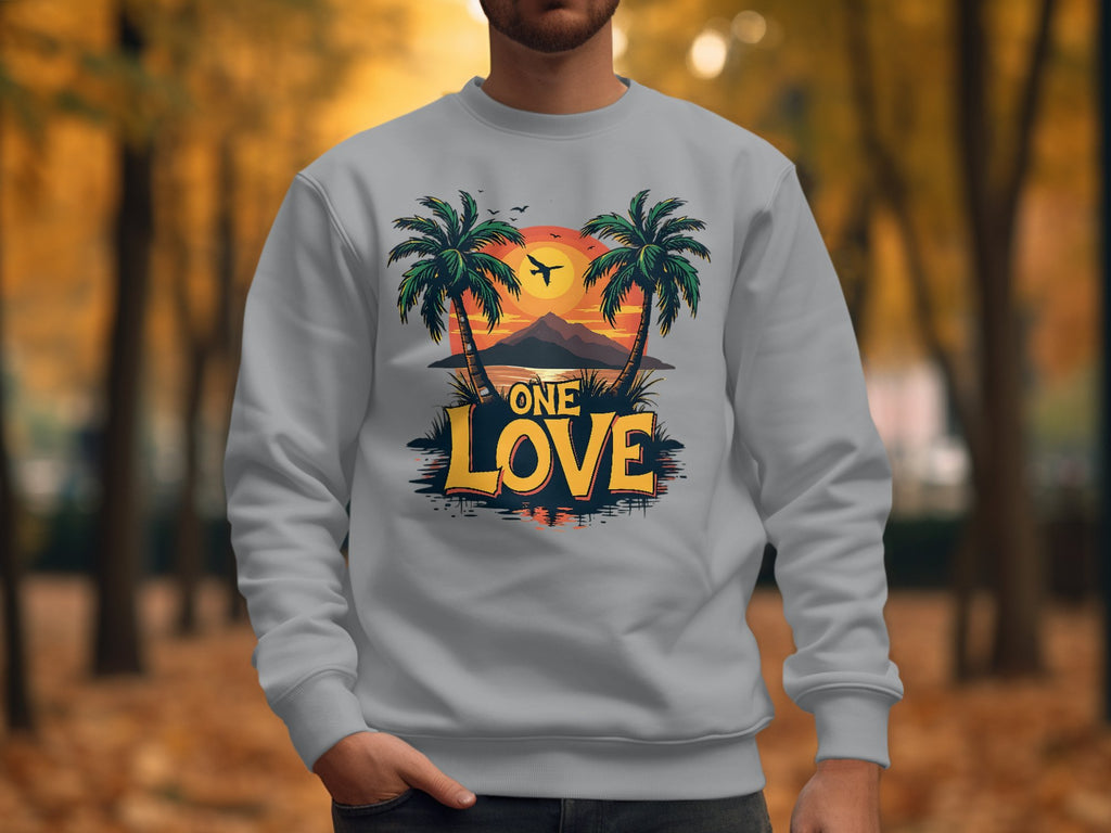 A person wearing the unisex-sized One Love Sweatshirt by Garment Graphics, featuring a tropical design of two palm trees framing a sunset over a mountain, with One Love text, stands against blurry autumn trees and fallen leaves.