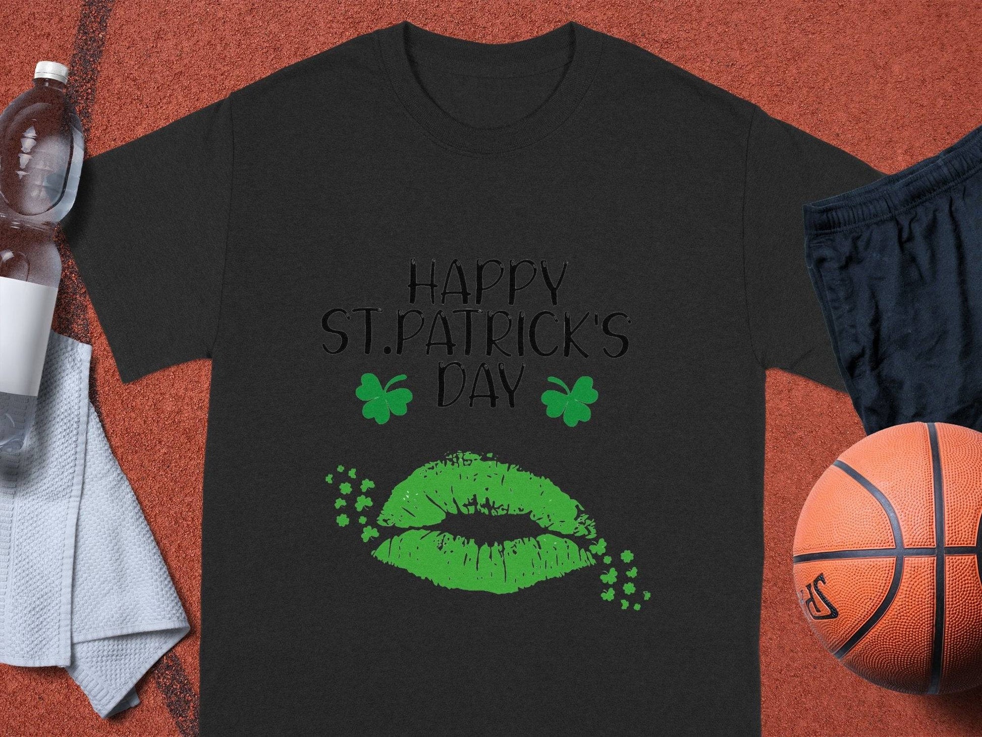 A Garment Graphics black graphic T-shirt displays Happy St. Patricks Day with green lips and clovers. Nearby lie a water bottle, white towel, black shorts, an orange basketball, all on a red surface.