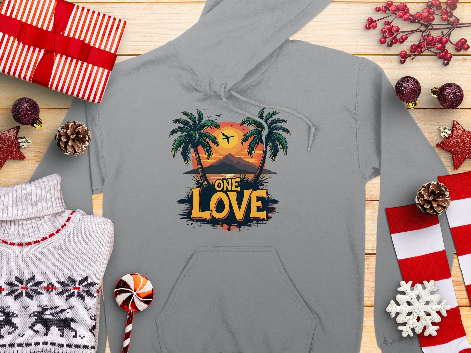 A Garment Graphics gray hoodie showcases a tropical sunset with palm trees, an airplane, and One Love, surrounded by Christmas items: wrapped gifts, red ornaments, pinecones, a cozy sweater, candy canes, and star decorations on a wooden surface.