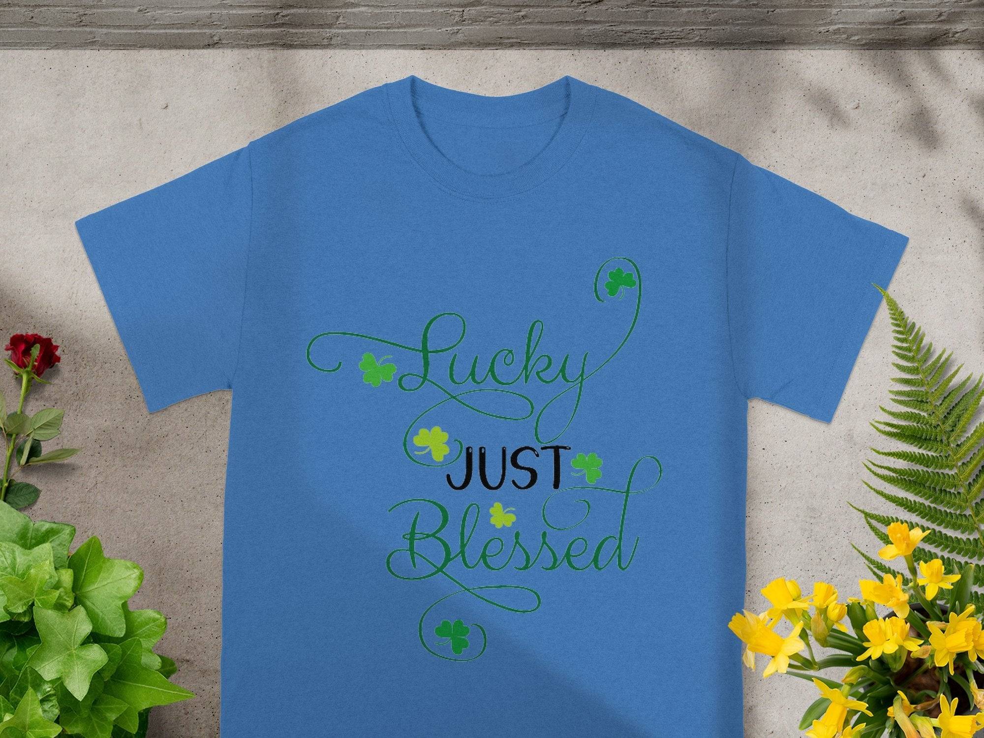A Royal Mail - 24 Tracked classic fit blue T-shirt rests on concrete, featuring Lucky Just Blessed in green script with shamrock designs. Its surrounded by potted plants with lush green leaves and vibrant yellow and red flowers.