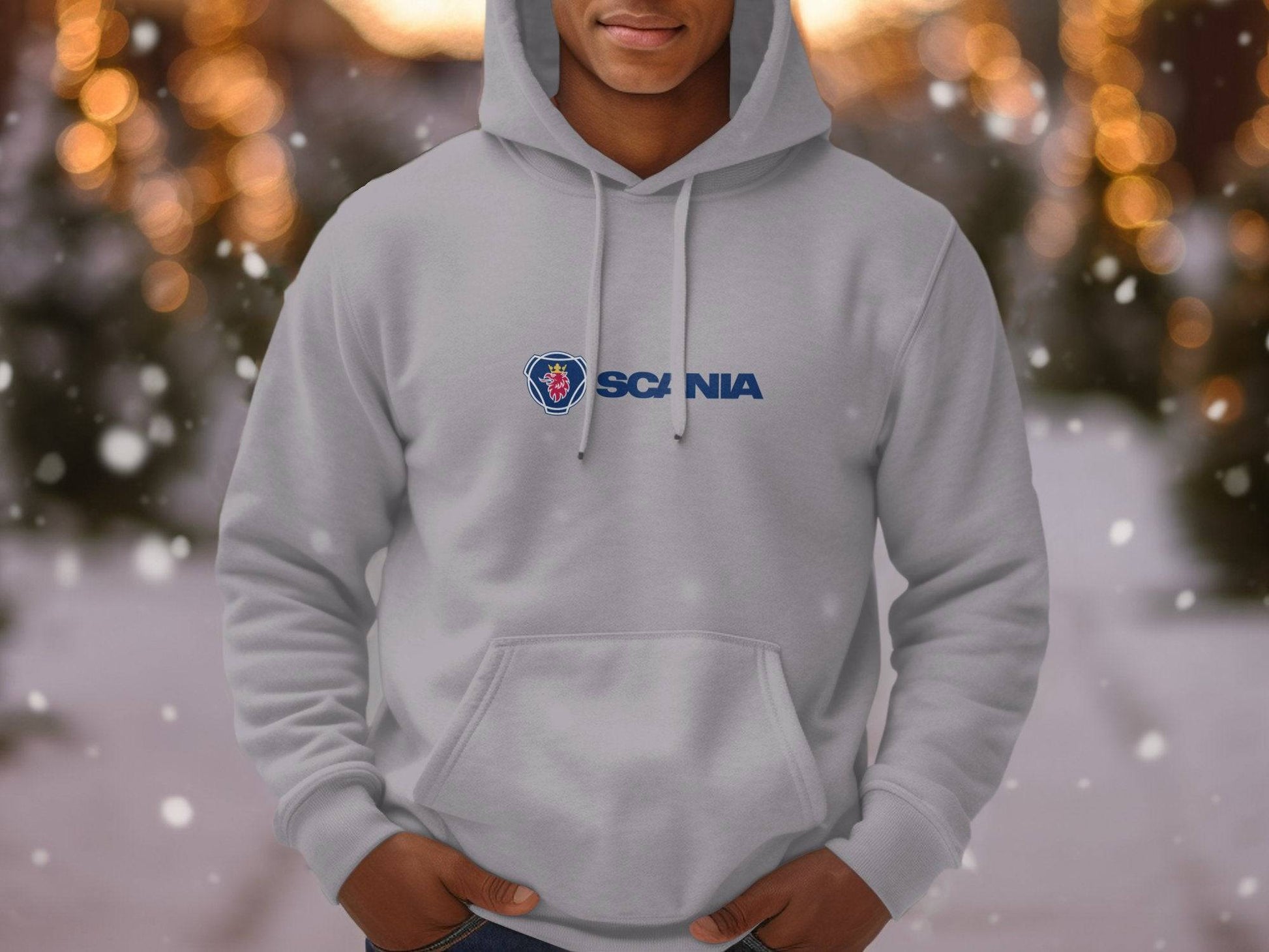 Wearing a light gray hoodie from Garment Graphics featuring the iconic Scania logo, an individual stands outdoors surrounded by blurred trees and snowflakes, capturing a wintry essence. This fashion piece exemplifies a perfect blend of style and functionality.