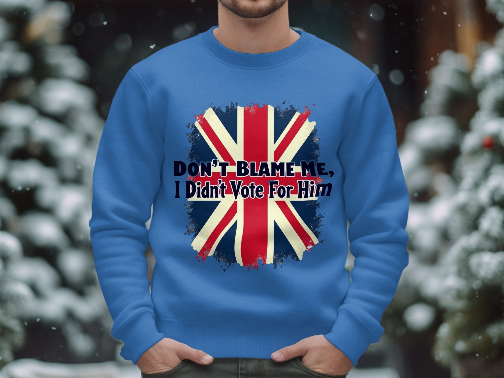 Wearing a cozy blue Garment Graphics sweatshirt with a bold British flag design and the text Dont blame me, I didnt vote for him, a person stands with snow-covered trees blurred in the background.